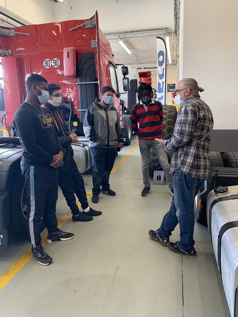 cours-en-atelier-POEC-Technopolys Un enseignant dispense un cours à des stagiaires au milieu de poids lourds dans un atelier.